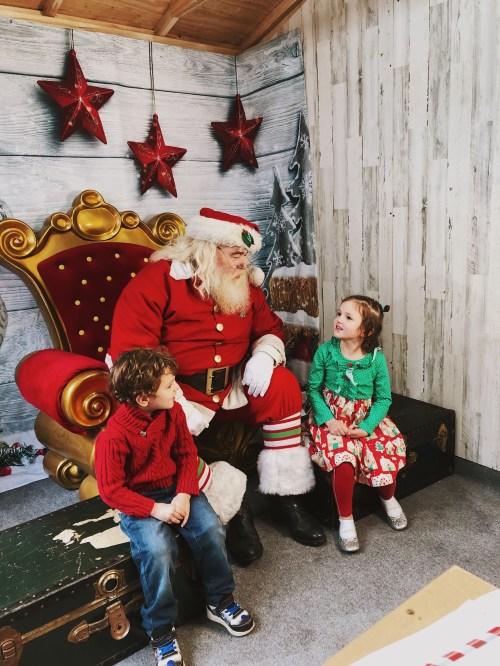 A jolly Santa sits on a throne with two smiling children, surrounded by festive decorations and stars.