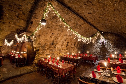 Cozy underground dining area with stone walls, festive lights, and tables set with red napkins and candles.
