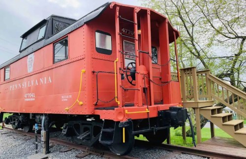 A vintage red caboose labeled "Pennsylvania 47704" next to a wooden staircase and greenery.