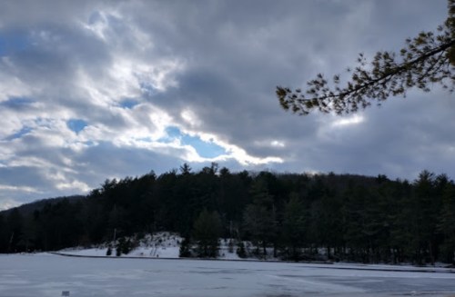 A snowy landscape with a frozen lake, surrounded by trees and a cloudy sky.