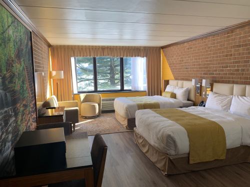 A cozy hotel room with two beds, a large window, and a seating area, featuring warm colors and a brick wall.