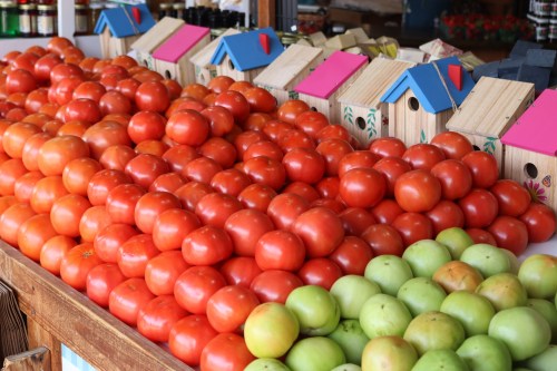 A vibrant display of red tomatoes and green apples at a market, with colorful wooden birdhouses in the background.