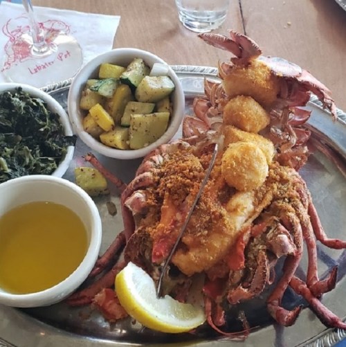 A plated lobster with stuffing, served with sautéed vegetables, greens, and a side of olive oil and lemon.
