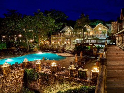 A beautifully lit lodge at night, featuring a pool surrounded by trees and cozy seating areas.
