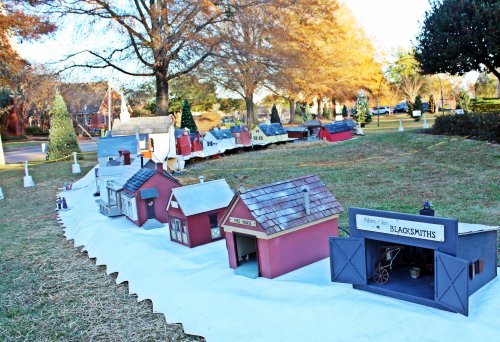 A miniature village display with various colorful buildings, set on a grassy area surrounded by trees and holiday decorations.