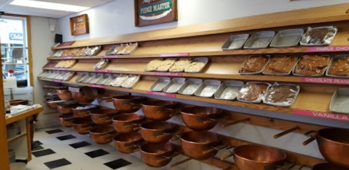 A display of various ice cream flavors in metal trays on wooden shelves, with copper mixing bowls below.