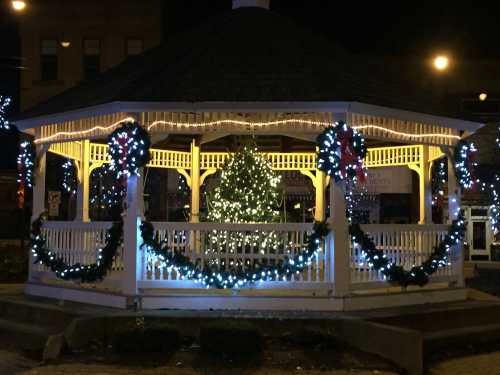 A decorated gazebo at night, adorned with lights and wreaths, featuring a glowing Christmas tree inside.