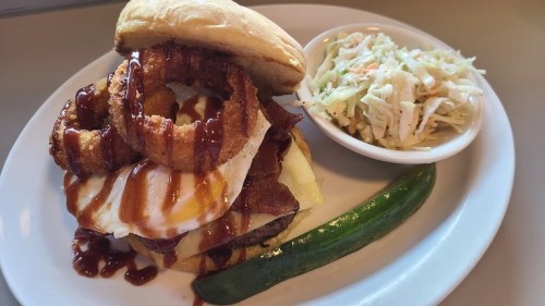 A hearty burger topped with onion rings, bacon, and barbecue sauce, served with coleslaw and a green pepper on the side.