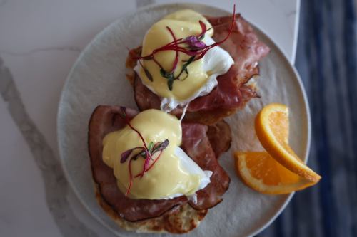 Two eggs Benedict on toasted English muffins with ham, topped with hollandaise sauce and microgreens, served with an orange slice.