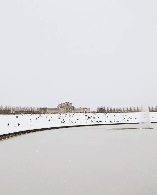 A snowy landscape with people walking on a frozen lake, surrounded by trees and a distant building.