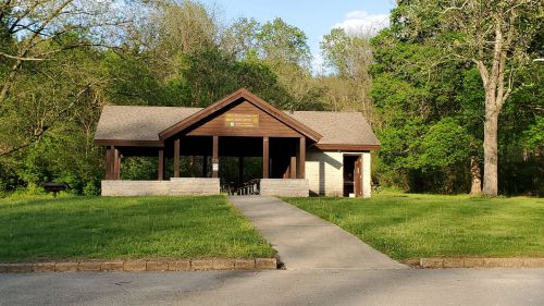 A wooden pavilion with a sloped roof, surrounded by trees and grass, with a concrete path leading to the entrance.