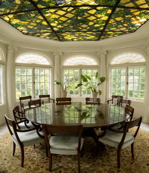 A round wooden dining table surrounded by chairs in a bright room with large windows and a stained glass ceiling.