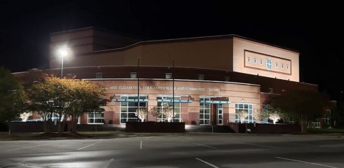 A well-lit community center building at night, featuring large windows and surrounding trees.