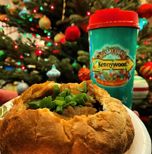 A festive scene featuring a filled bread bowl topped with green onions, with a holiday-themed cup and a decorated tree in the background.