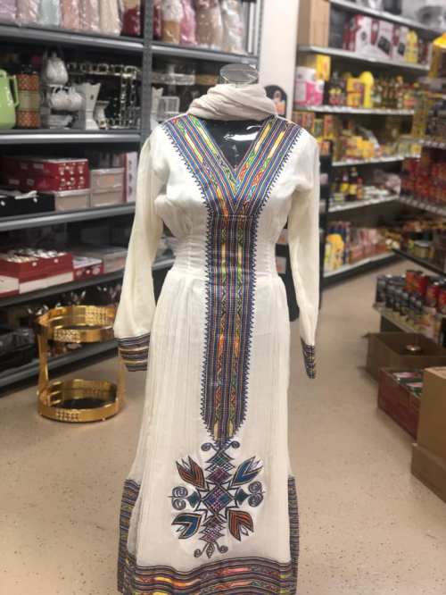 A white dress with colorful embroidery displayed on a mannequin in a store aisle filled with various products.