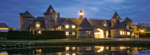A stone castle with turrets, illuminated windows, and a reflective pond at dusk.