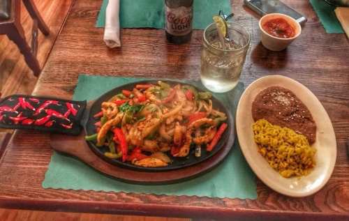 Sizzling fajitas with chicken, peppers, and onions on a wooden platter, served with rice and beans, plus a drink.