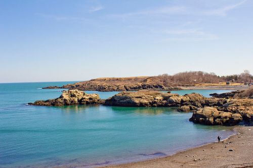 A serene coastal scene with rocky formations and calm blue waters under a clear sky, with a person walking along the shore.