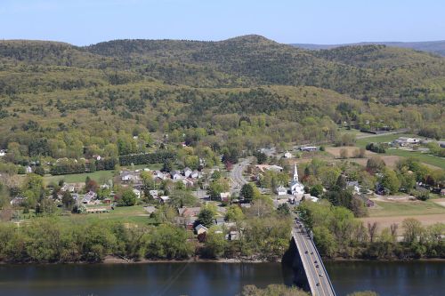 Aerial view of a lush green landscape with a river, a bridge, and a small village surrounded by hills.