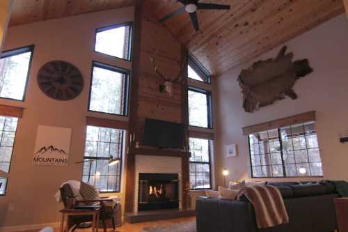 Cozy living room with high ceilings, large windows, a fireplace, and rustic decor featuring wood and antlers.