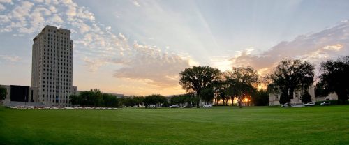 A panoramic view of a grassy field at sunset, with a tall building and trees silhouetted against the colorful sky.