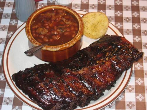 A plate featuring a large portion of glazed ribs, a bowl of baked beans, and a cornbread muffin on a checkered tablecloth.