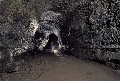 A person walks through a dark, rocky cave with a light, revealing textured walls and a sandy floor.