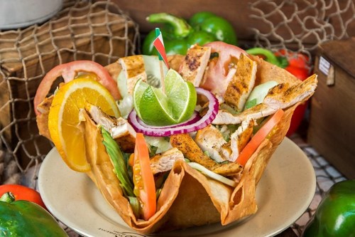 A colorful taco salad bowl filled with grilled chicken, fresh vegetables, and garnished with lime and orange slices.