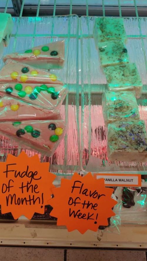 Display of colorful fudge varieties with signs reading "Fudge of the Month!" and "Flavor of the Week!" in a shop.