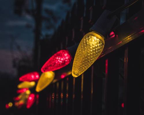 Colorful holiday lights in red and yellow glow against a dark background, illuminating a fence.