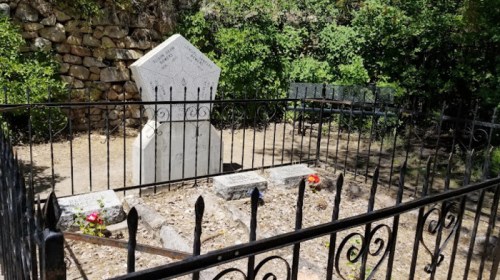 A fenced grave site with a white tombstone and flowers, surrounded by greenery and stone walls.