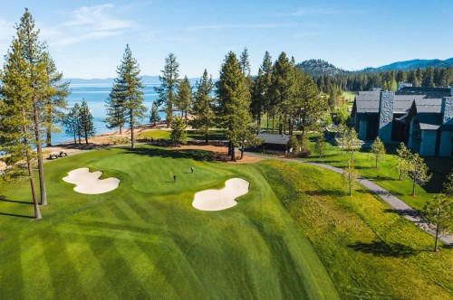 Aerial view of a golf course by a lake, surrounded by trees and mountains, with a clubhouse in the background.