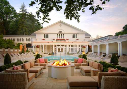 A luxurious outdoor lounge area with a fire pit, surrounded by seating and a pool, set against a grand white building.