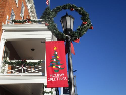 A festive street scene featuring a holiday banner and a decorated lamppost with a wreath and ornaments against a blue sky.