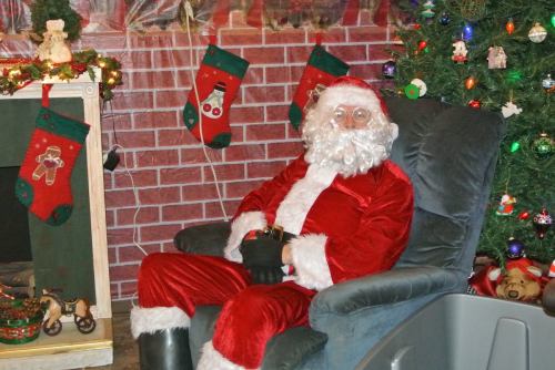 A person dressed as Santa Claus sits in a chair, surrounded by Christmas stockings and a decorated tree.