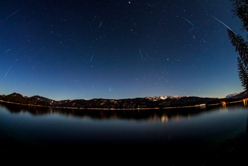 A serene night sky filled with stars and meteor trails above a calm lake, surrounded by mountains.