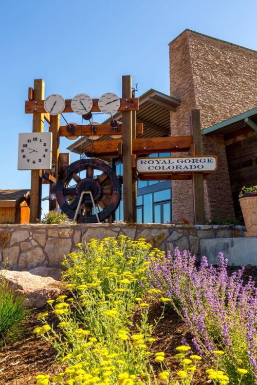 Sign for Royal Gorge, Colorado, featuring clocks and a ship's wheel, surrounded by colorful flowers.