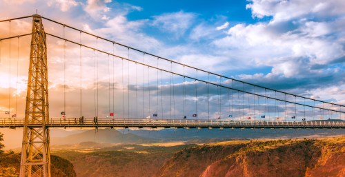 A suspension bridge spans a canyon, with flags along its length and a colorful sunset in the background.