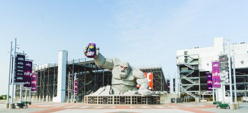 A large stone gorilla statue holding a can, with banners and a stadium in the background under a clear sky.