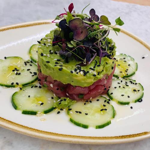A layered dish featuring avocado and tuna, garnished with microgreens and served on a plate with cucumber slices.