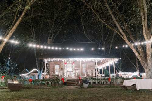 A charming wooden house decorated with string lights and festive wreaths, surrounded by trees at night.