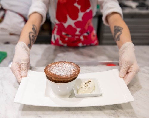A person in gloves presents a chocolate soufflé with powdered sugar and a scoop of ice cream on a white plate.