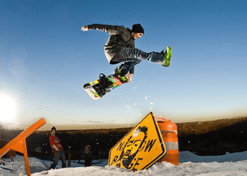 A snowboarder jumps over a construction sign, with snow and a sunset backdrop, while another person watches nearby.