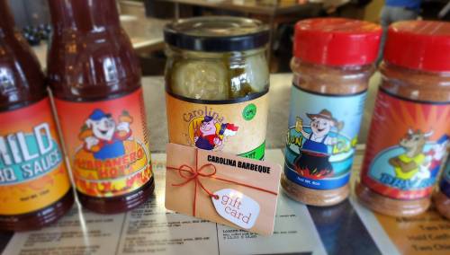 A gift card sits on a table surrounded by barbecue sauces and spices, featuring colorful labels and a jar of pickles.