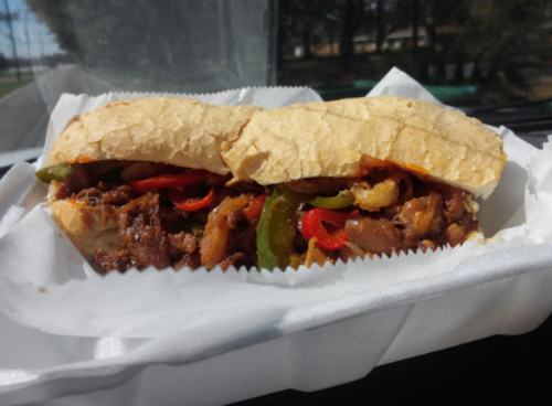 A sandwich filled with seasoned meat, peppers, and onions, served in a soft hoagie roll on a white paper tray.