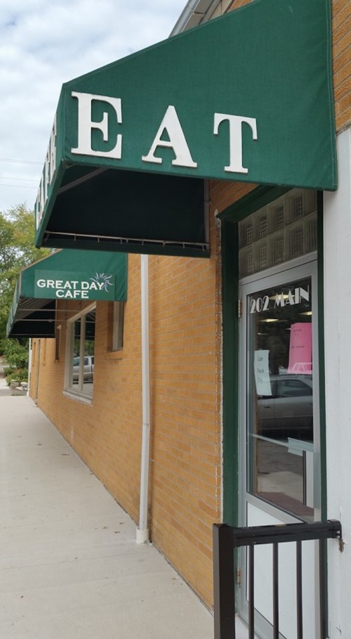 Exterior view of a cafe with green awnings reading "EAT" and "GREAT DAY CAFE" on a brick building.