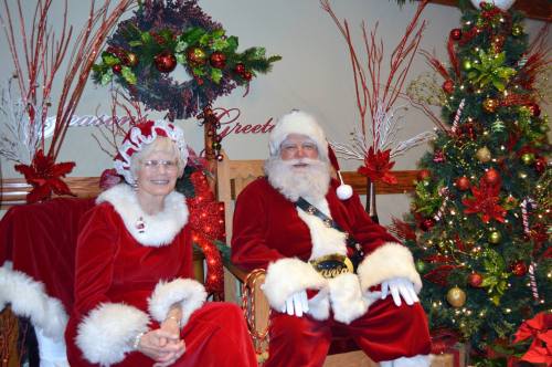 A cheerful Santa Claus and a woman in a festive outfit sit together, surrounded by holiday decorations and a Christmas tree.