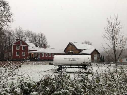 A snowy landscape featuring a red farmhouse, a barn, and a white maple syrup wagon in the foreground.