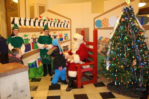 Santa Claus sits in a chair with children nearby, surrounded by festive decorations and a Christmas tree.