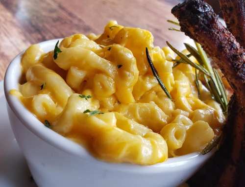 Creamy macaroni and cheese in a white bowl, garnished with herbs, alongside a crispy piece of roasted meat.